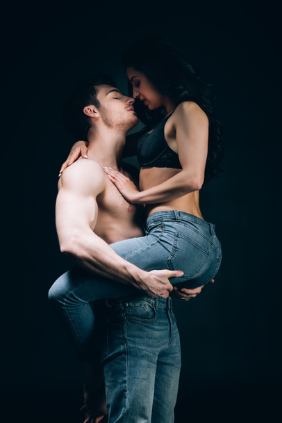 sexy man in jeans with muscular torso holding girlfriend in hands isolated on black - Fotoğraf, Görsel