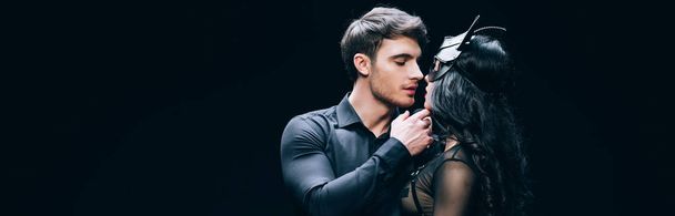 Panoramaaufnahme von jungen Mann küsst sexy brünette Frau in bdsm Kostüm isoliert auf schwarz - Foto, Bild