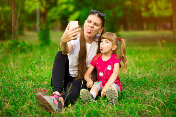 Автопортрет сумасшедшей, мать и дочь показывают язык, ребенок делает селфи на мобильном телефоне на фоне природы, проводит выходные, женский день вместе
 - Фото, изображение
