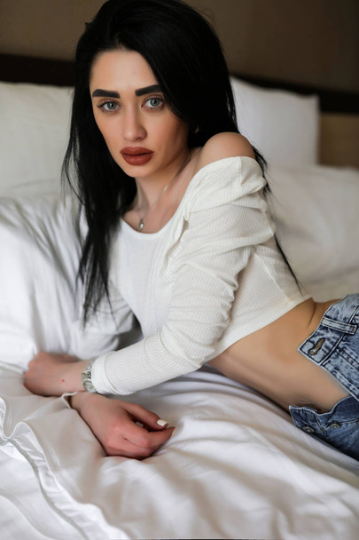 Beauty portrait of young brunette model lying in her bed alone. Green eyes, big lips, makeup, stylish hair. Wearing fancy white top and jeans. Soft sheets and pillows make the room cozy. - Photo, Image