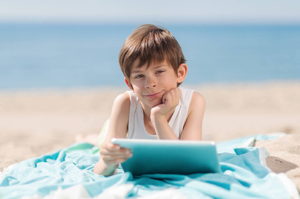 Jongen met Tablet liggend op het strand een zonnige dag kijken naar camera - Foto, afbeelding