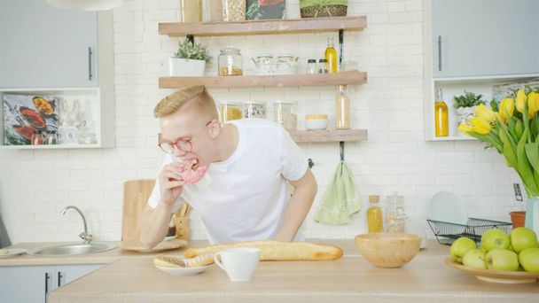 Blonde man is eating donut for breakfast at the kitchen at home - Фото, зображення