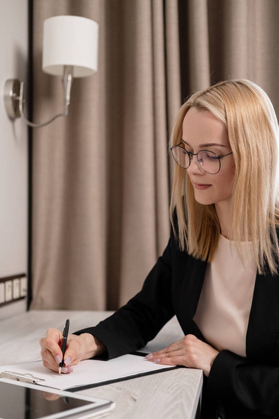 Attractive blonde woman in glasses and a business suit working on a tablet, talking on the phone and taking notes in a notebook. Hotel room, table by the window. - Foto, Imagem