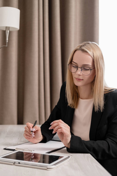 Attractive blonde woman in glasses and a business suit working on a tablet, talking on the phone and taking notes in a notebook. Hotel room, table by the window. - Zdjęcie, obraz