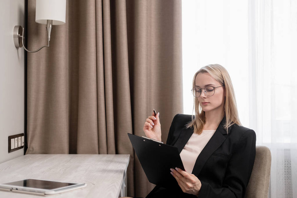 Attractive blonde woman in glasses and a business suit working on a tablet, talking on the phone and taking notes in a notebook. Hotel room, table by the window. - Valokuva, kuva