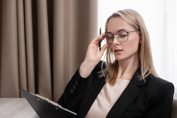 Attractive blonde woman in glasses and a business suit working on a tablet, talking on the phone and taking notes in a notebook. Hotel room, table by the window. - Фото, изображение