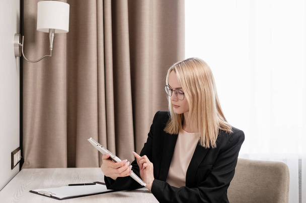 Attractive blonde woman in glasses and a business suit working on a tablet, talking on the phone and taking notes in a notebook. Hotel room, table by the window. - Фото, изображение