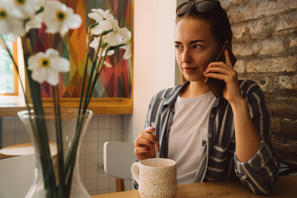 Joven mujer bonita sentada en un café y hablando por teléfono. La chica bebe café caliente
 - Foto, imagen