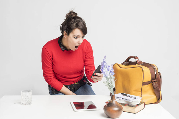 Vorderseite einer schönen lateinischen Frau, die auf einem Schemel sitzt, während sie ein Mobiltelefon benutzt und im Büro vor weißem Hintergrund überrascht - Foto, Bild