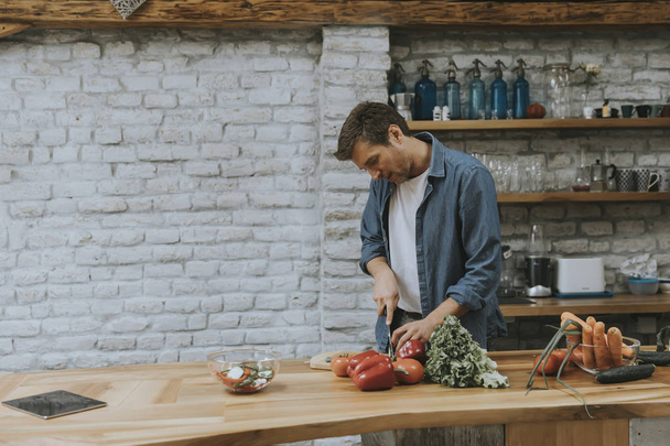 Giovane uomo che taglia verdure fresche nella cucina rustica e prepara pasti sani
 - Foto, immagini