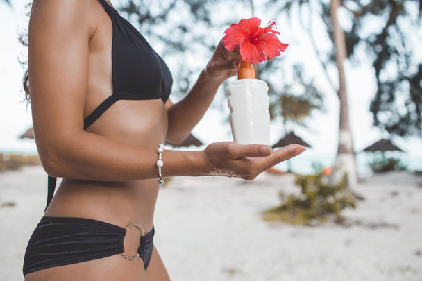 Tanned young woman with perfect body with tattoo on her hand and red flower is holding jar with sunscreen on the beach. Beauty and fashion concept. - 写真・画像
