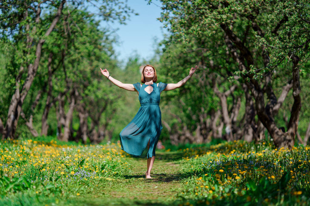 Повнометражне фото жінки з закритими очима і піднятими руками в довгій зеленій сукні робить йогу в лісі
 - Фото, зображення