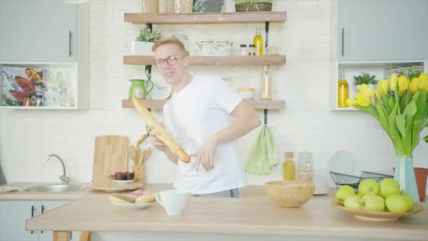 Young man is goofing around and singing with bread instead of microphone at the kitchen - Materiał filmowy, wideo