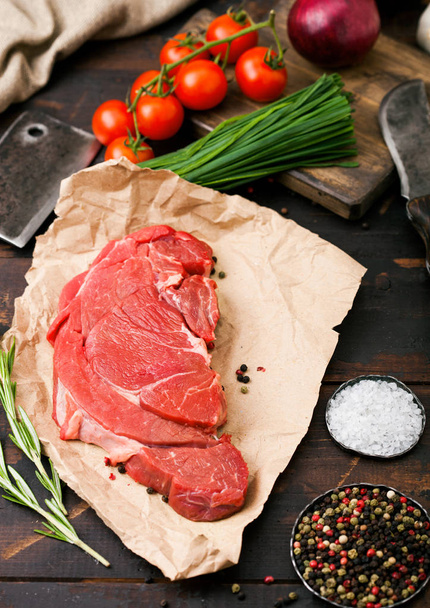 Fresh raw organic slice of braising steak fillet on butchers paper with fork and knife on dark wooden background. Red onion, tomatoes with salt and pepper. - Foto, Bild
