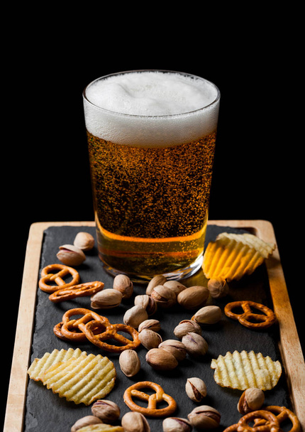 Glass of lager beer with snack on stone board on black background. Pistachios and pretzel with potato crisps.  - Foto, afbeelding