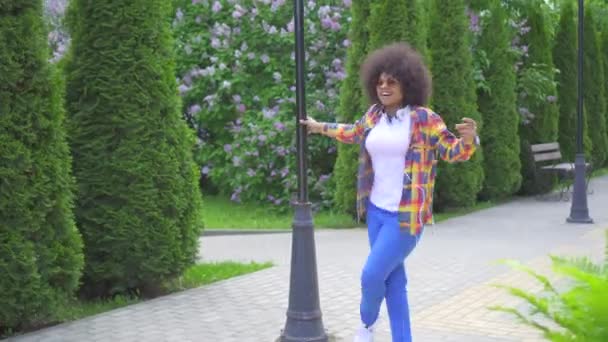cheerful happy african woman with an afro hairstyle with headphones walking down the street dancing and singing - Materiał filmowy, wideo