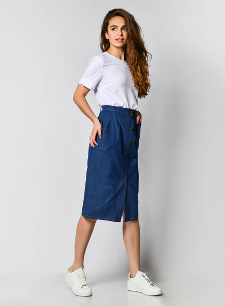 Full-length photo of a young brunette girl with long hair in a white blouse and blue skirt. on a light background - Foto, Imagen