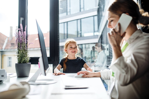 Una mujer de negocios con una hija pequeña sentada en una oficina, trabajando
. - Foto, Imagen
