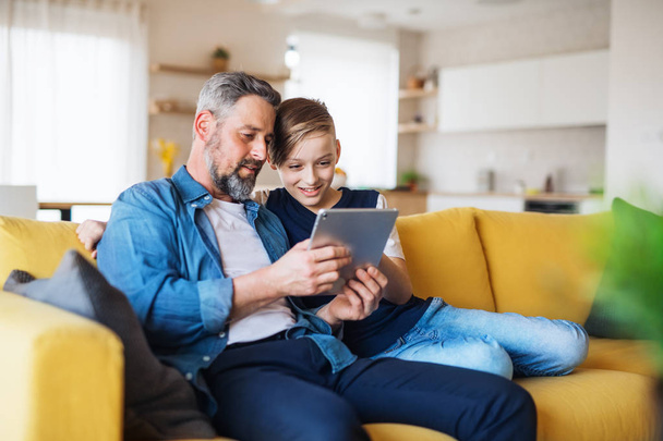Mature father with small son sitting on sofa indoors, using tablet. - Foto, Bild