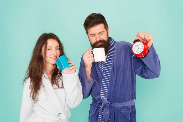 Man with beard and sleepy woman enjoy morning coffee or tea. Time to wake up and have nice day. Guy husband hold coffee and clock. Every morning begins with coffee. Couple in bathrobes with mugs - Zdjęcie, obraz