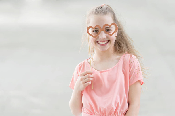 The goggles are accessory for her face. Little child looking happy through goggles props. Funny small girl with heart shaped goggles on stick. Cute kid smiling with fancy party googles, copy space - Фото, зображення