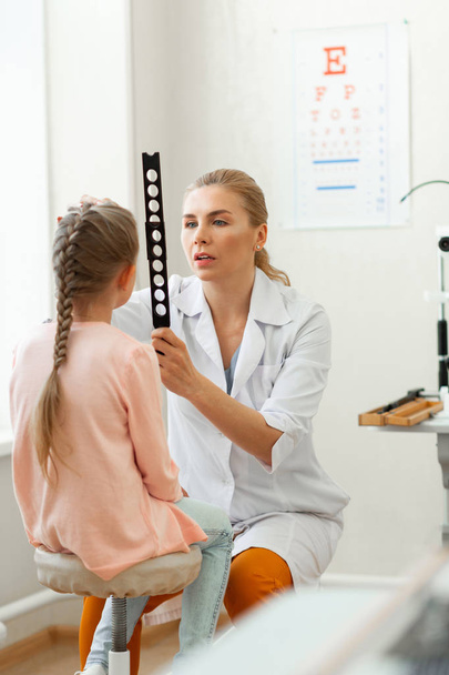 Exploring eyesight. Blonde caring doctor working with little girl while observing her eyes through measuring tool - Fotoğraf, Görsel