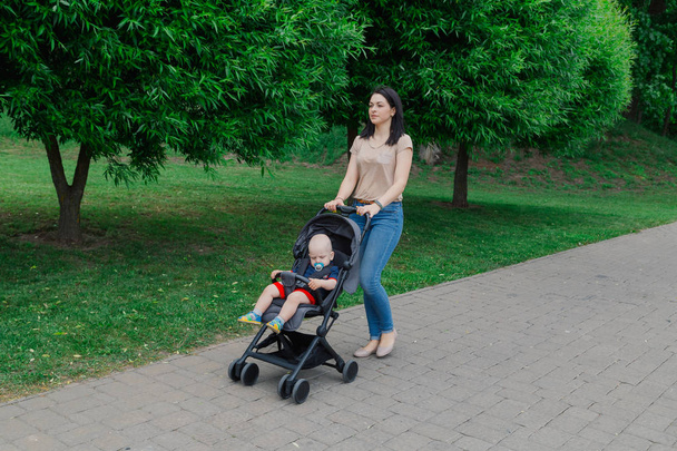 Hermosa mujer con un niño en un cochecito pasea por un parque de verano
. - Foto, imagen