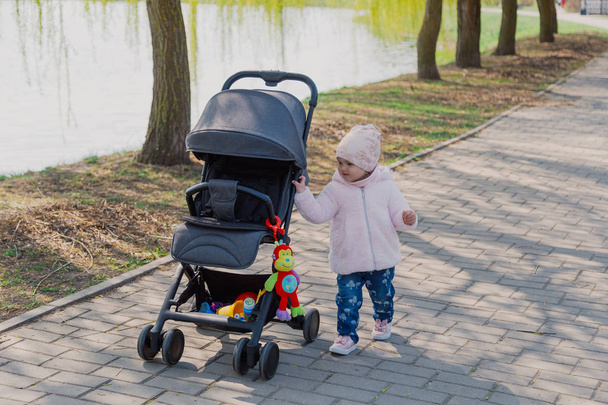 Uma menina está rolando carruagem de bebê no parque. Criança no parque brincando com carrinho
. - Foto, Imagem