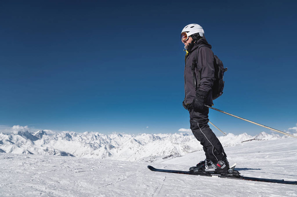 Ένας μουσάτος αρσενικός σκιέρ με κράνος και μάσκα του σκι στέκεται πάνω σε σκι στο φόντο των χιονισμένα βουνά και του γαλάζιου ουρανού. Αθλητής σε μαύρο κοστούμι - Φωτογραφία, εικόνα