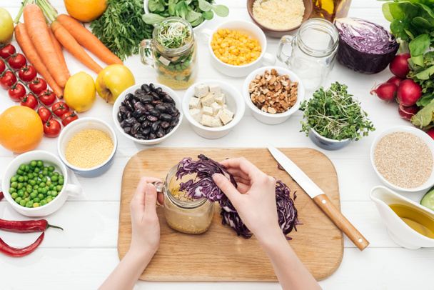 cropped view of woman adding cut red cabbage in jar with salad on wooden white table  - Foto, Imagem