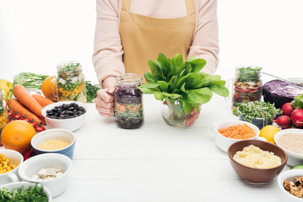 bijgesneden beeld van de vrouw in schort houden glazen pot met salade en groene bladeren op houten tafel geïsoleerd op wit - Foto, afbeelding