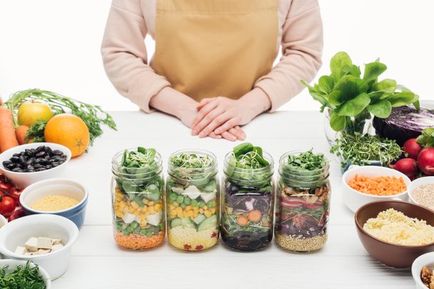 partial view of woman in apron standing near glass jars with salad on wooden table isolated on white - Photo, Image