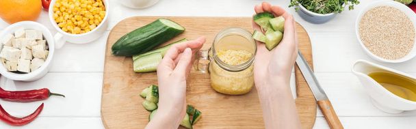 cropped view of woman adding chopped cucumber to couscous in glass jar on wooden chopping board, panoramic shot - Photo, Image