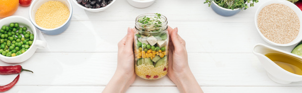 cropped view of woman holding glass jar with fresh salad on wooden white table, panoramic shot - Photo, Image