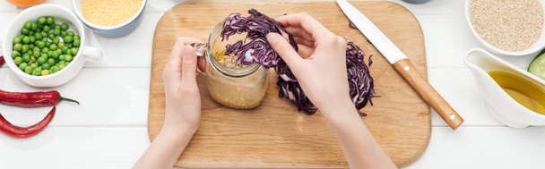 vista cortada da mulher adicionando o repolho vermelho cortado no jarro com salada na mesa branca de madeira, tiro panorâmico
 - Foto, Imagem