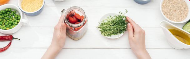 bijgesneden beeld van vrouw Holding jar met salade en groene spruiten op houten witte tafel, panoramische shot - Foto, afbeelding