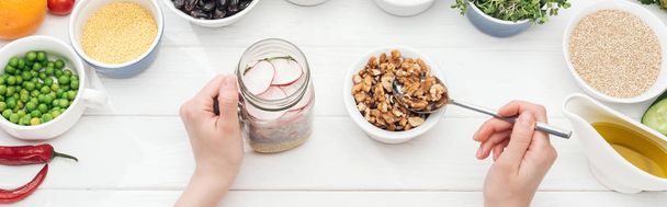 vista recortada de mujer añadiendo nueces en tarro con ensalada sobre mesa blanca de madera, plano panorámico
 - Foto, imagen