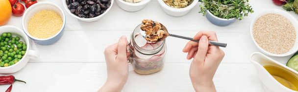 cropped view of woman adding nuts in glass jar with salad on wooden white table, panoramic shot - Photo, Image