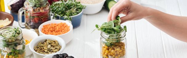 cropped view of woman adding greens in glass jar on wooden white table, panoramic shot - Photo, Image