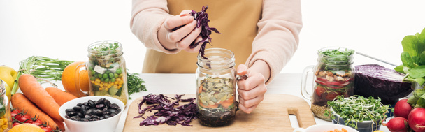cropped view of woman in apron adding red cabbage in glass jar on wooden table isolated on white, panoramic shot - Foto, Imagem