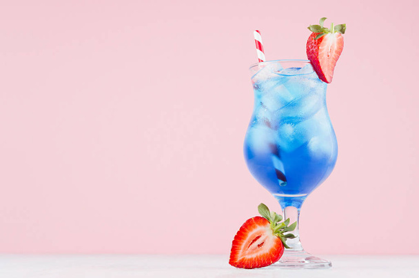 Fresh alcohol blue cocktail with curacao liquor, strawberry slice, ice cubes, red striped straw in misted glass on pink background. - Photo, image
