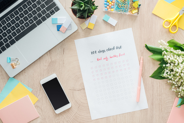 bovenaanzicht van 100 dagen checklist met smartphone, laptop en briefpapier op houten tafel - Foto, afbeelding