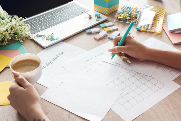 cropped view of woman holding cup of coffee in hand, sitting behind wooden table with laptop and stationery, writing in paper planners - Photo, Image
