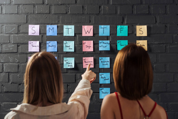 back view of two women standing near colorful sticker notes and pointing with finger on black brick wall - Photo, Image