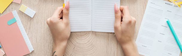 cropped view of woman holding notepad in hands over wooden table with weekly list and stationery - Photo, Image