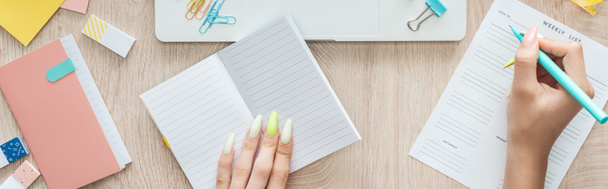 Ausgeschnittene Ansicht einer Frau, die Notizen in der Wochenliste macht, hinter einem Holztisch mit Notizblock und Schreibwaren sitzt - Foto, Bild
