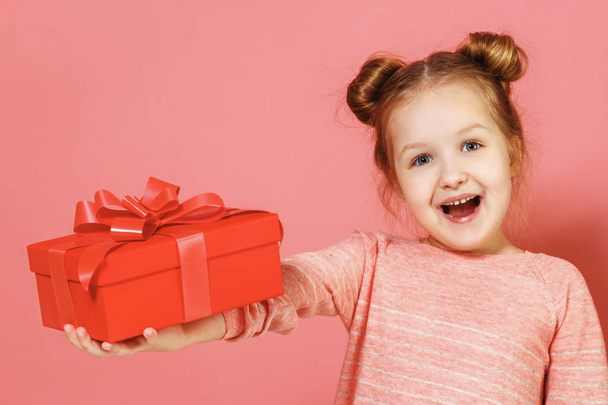 Closeup portrait of a cute little girl with hair buns on pink background. The child holds out a box with a gift. - Foto, imagen