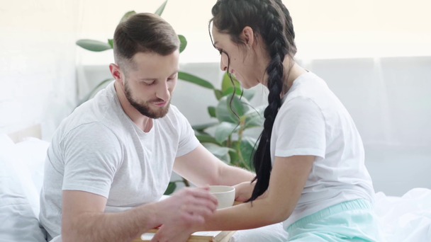 cheerful man using smartphone while woman bringing breakfast on tray, boyfriend kissing girlfriend and eating in bedroom  - Video, Çekim