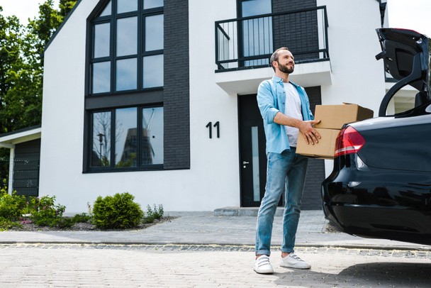 ευτυχισμένος άνθρωπος κρατώντας κουτιά κοντά στο αυτοκίνητο και μοντέρνο σπίτι  - Φωτογραφία, εικόνα
