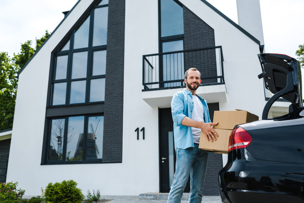 низький кут зору веселий чоловік тримає коробки біля автомобіля і сучасного будинку
  - Фото, зображення
